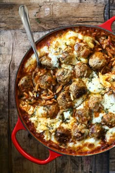 a red pot filled with meatballs and cheese on top of a wooden table next to a spoon