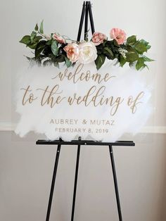 a welcome sign with flowers and greenery is displayed on a easel in front of a white wall