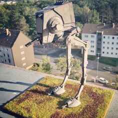 a statue of a robot on top of a grass covered field in front of buildings