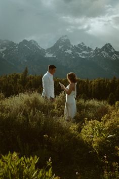 a man and woman standing on top of a lush green field next to mountains in the background