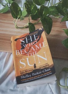 a book sitting on top of a bed next to a green leafy plant with the title she who came the sun