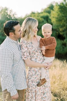 a man and woman holding a baby while standing in tall grass with trees in the background