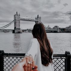 a man and woman holding hands while standing next to the water in front of a bridge