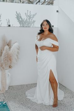 a woman in a white dress standing next to a plant and posing for the camera