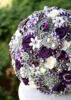 a bridal bouquet with purple and white flowers on top of a stone pedestal in front of some bushes