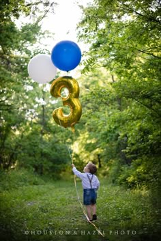 a young boy is holding two balloons in the air while walking down a path through some trees