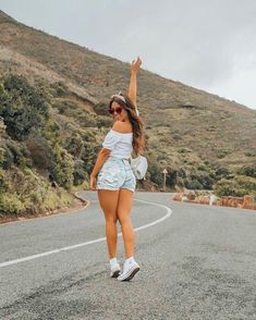 a woman in white shirt and blue shorts standing on the side of road with her arms up