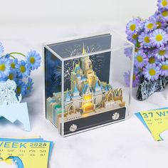 a clear box with a castle in it surrounded by purple flowers and blue tags on the side