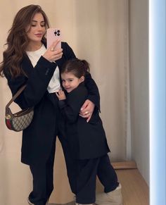 a woman taking a selfie with her daughter in front of a mirror while she is wearing a black blazer and white shirt