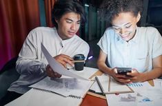 two people sitting at a table looking at their cell phones and paper work on them