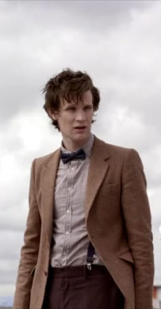 a man in a brown jacket and bow tie standing on the beach with his hands in his pockets