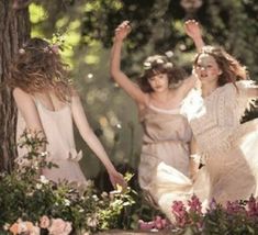 three women in white dresses are walking through the woods with their arms up and hands raised