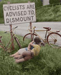 two people laying in the grass next to bicycles and a sign that says cyclists are advised to dismount