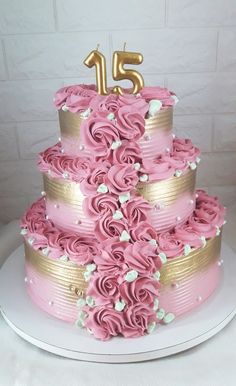 a pink and gold birthday cake with flowers on the bottom tier is displayed in front of a white brick wall