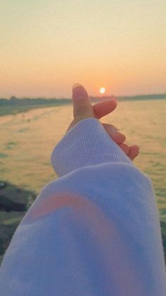 someone is holding their hand out to the water at sunset or sunrise, with the sun setting in the distance