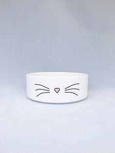 a white bowl with a cat's face painted on the front and side, sitting on a gray surface