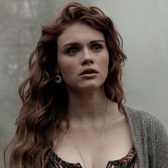 a woman with long red hair wearing a gray cardigan and silver necklace standing in front of a wall