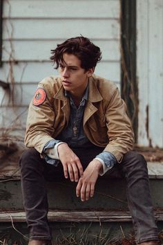 a man sitting on top of a wooden bench next to a white wall and wearing a brown jacket