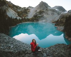 a woman sitting on top of a rock next to a blue lake in the mountains