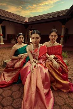 three women sitting on the ground wearing different colored saris and necklaces, all looking at the camera