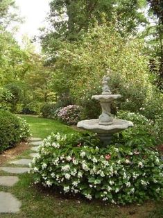 a stone fountain in the middle of a garden with white flowers and trees around it