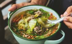 two hands holding a bowl of soup with meat and vegetables in it, while another hand holds a spoon