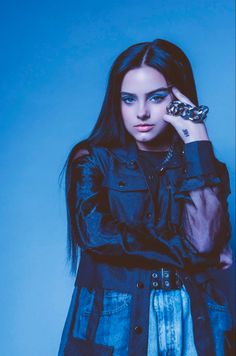 a woman with long dark hair wearing a black jacket and jeans is holding her hand up to her face