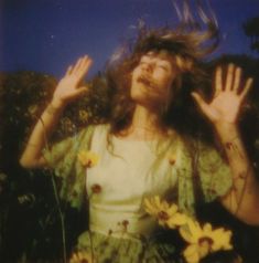 a woman with long hair standing in front of flowers and holding her hands up to the sky