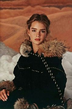 a woman sitting in the snow wearing a black coat with fur collar and cuffs on her shoulders