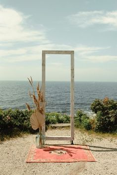 a wooden frame sitting on top of a sandy beach next to the ocean with a vase filled with flowers