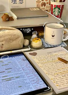 an open book on a desk next to a tablet and coffee cup with a pen