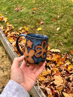 a hand holding a blue and brown coffee mug in front of leaves on the ground