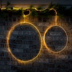 two circular lights are hanging on a brick wall with pine cones and evergreen branches in the background