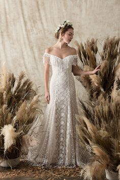 a woman in a white dress standing next to some tall grass and flowers with her hands on her hips