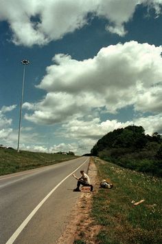 a man kneeling down on the side of a road next to a street light with clouds in the sky