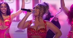 two women in red and pink outfits singing into microphones while another woman holds her arm up