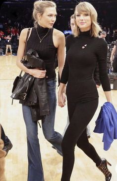 two women walking on a basketball court holding hands and looking at each other with people in the background