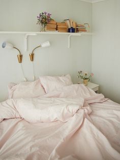 an unmade bed with pink sheets and pillows in a white room next to bookshelves