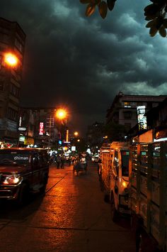 cars and trucks are parked on the street in front of buildings at night with dark clouds overhead