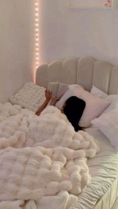 a woman laying on top of a white bed covered in blankets and pillows with lights behind her