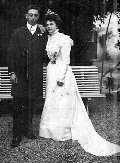 an old black and white photo of a man and woman in formal wear standing next to each other on a bench