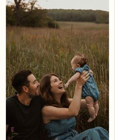 a man and woman holding a baby in their arms while sitting on the ground with tall grass behind them