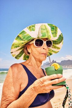 an older woman wearing sunglasses and a hat drinking from a green cup on the beach