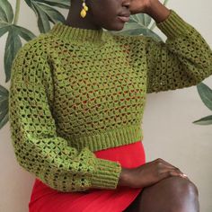 a woman wearing a green sweater and red skirt sitting on a chair in front of a plant