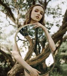 a woman is holding a mirror in front of her face while standing next to a tree