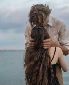 a man and woman standing next to each other near the ocean with long hair in their hands