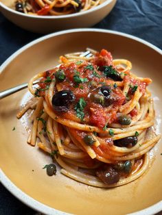 a plate of spaghetti with olives and tomato sauce on it, next to a bowl of pasta