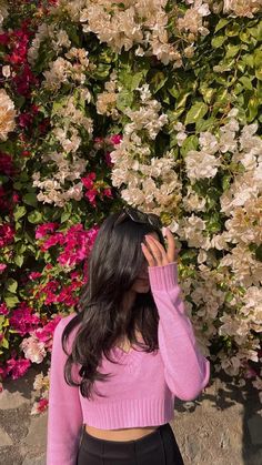 a woman wearing a pink sweater and black skirt standing in front of flowers with her hands on her head