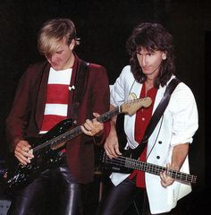 two young men are playing guitars together in the same room, one is wearing a white shirt and black leather pants