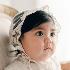 Baby girl with dark hair wearing the Penelope Lace Bonnet and matching baptism dress gazes away to the side of the camera. Elegant Lace Patchwork Tulle, Scalloped Lace Tulle Fabric, Lace Veil With Lace Trim For Ceremonies, Elegant Lace Veil With Lace Trim, Elegant Tulle Lace With Lace Trim, Lace Ceremony Veil With Lace Trim, Ceremony Lace Veil With Lace Trim, Ceremony Veil With Lace Trim, Fitted White Bonnet With Lace Trim